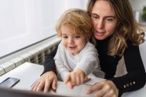 Femme employée à distance tenant un curieux petit enfant à genoux assis à table et travaillant avec un ordinateur portable à la maison — Photo de stock