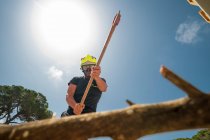 Снизу пожарный в защитной форме режет ветку топором в лесу на фоне голубого неба — стоковое фото