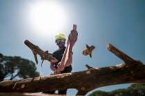 Von unten der Feuerwehrmann in Schutzuniform schneidet Ast mit Axt im Holz vor blauem Himmel — Stockfoto