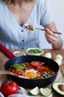 Donna irriconoscibile in abito seduto a tavola con vari ingredienti e godendo di shakshuka fresco con uova e verdure — Foto stock