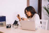 Молодая женщина-фотограф сидит за столом с ноутбуком и фотокамерой с проектором и работает со старыми слайдами на домашнем рабочем месте — стоковое фото