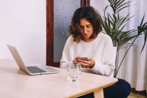 Side view of focused young female freelancer in casual clothes checking messages on smartphone while sitting at table with laptop and working on project at home — Stock Photo