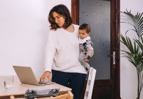 Young busy female in casual wear with little kid on hand standing at table and checking email on laptop while working online at home — Stock Photo