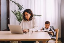 Young female freelancer in casual clothes sitting at table and working on project with laptop while little child sitting nearby with book — Stock Photo