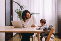 Young female freelancer in casual clothes sitting at table and working on project with laptop while little child sitting nearby with book — Stock Photo
