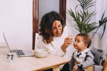 Modern young female remote worker feeding little child while sitting at table with laptop and working on online project — Stock Photo