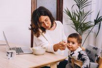 Moderna giovane lavoratrice remota che allatta il bambino mentre siede a tavola con il computer portatile e lavora al progetto online — Foto stock