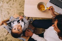 From above modern young female remote worker feeding little child while sitting at table with laptop and working on online project — Stock Photo