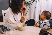 Modern young female remote worker feeding little child and discussing business issues on smartphone while sitting at table with laptop and working on online project — Stock Photo