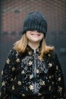 Смешной анонимный ребенок в теплой куртке и вязаной кепке, покрывающий половину лица, стоящего на улице и улыбающегося — стоковое фото