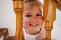Funny cute girl in white shirt placing head between wooden railings of stairs looking at camera — Stock Photo