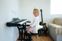 Side view of schoolgirl in fluffy skirt sitting at synthesizer and preparing for music class looking at camera — Stock Photo