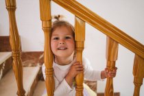 Funny cute girl in white shirt placing head between wooden railings of stairs looking at camera — Stock Photo