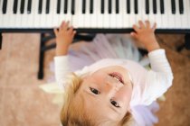 Von oben ein charmantes blondes kleines Mädchen, das fröhlich in die Kamera lacht, während es zu Hause Synthesizer spielt — Stockfoto