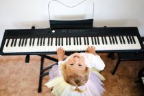 D'en haut de charmante petite fille blonde riant vivement à la caméra tout en jouant synthétiseur à la maison — Photo de stock