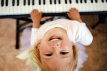 From above of charming blond little girl laughing brightly at camera while playing synthesizer at home — Stock Photo