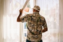 Visão traseira do homem irreconhecível em cap e camisa cortinas de luz de abertura de janela e olhando no quintal ensolarado — Fotografia de Stock