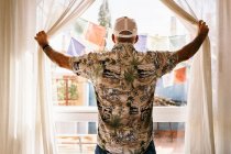 Visão traseira do homem irreconhecível em cap e camisa cortinas de luz de abertura de janela e olhando no quintal ensolarado — Fotografia de Stock