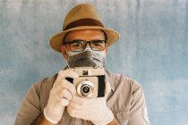 Чоловік середнього віку в латексних рукавичках і медичній масці фотографується з ретро-камерою в легкій студії, дивлячись на камеру — стокове фото