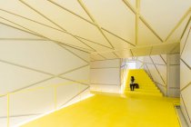 Женщина в повседневной одежде сидит в футуристическом коридоре желтой лестницы с геометрическими стенами и потолком — стоковое фото