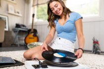 Feliz joven melomán femenino en traje casual sentado en la alfombra en el suelo cerca de la guitarra y escuchar música con disco de vinilo y tocadiscos mientras descansa en casa - foto de stock