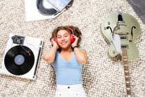 From above of happy young female looking at camera in casual wear lying on floor near guitar and listening to music with headphones and vinyl player while enjoying free time at home — Stock Photo