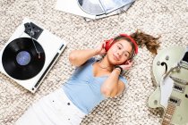 Зверху щаслива молода жінка дивиться в повсякденному одязі лежить на підлозі біля гітари і слухає музику з навушниками і вініловим плеєром, насолоджуючись вільним часом вдома — стокове фото