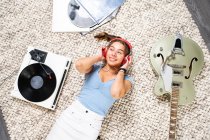 From above of happy young female looking away in casual wear lying on floor near guitar and listening to music with headphones and vinyl player while enjoying free time at home — Stock Photo