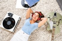 Сверху счастливая молодая женщина смотрит на камеру в повседневной одежде лежащей на полу рядом с гитарой и слушает музыку с наушниками и виниловым проигрывателем, наслаждаясь свободным временем дома — стоковое фото