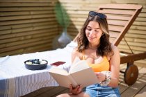 Cheerful female in summer top and shorts sitting near deck chair on wooden veranda and enjoying story while smiling and looking away — Stock Photo