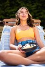 Весела жінка в літньому одязі сидить на стільці з стиглими фруктами та ягодами під час охолодження на дерев'яній терасі та озирається — стокове фото