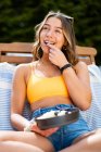 Веселая женщина в летнее время носить сидя на шезлонге с спелыми фруктами и ягодами, охлаждая на деревянной террасе и глядя в сторону — стоковое фото