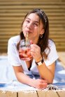 Joven mujer alegre en ropa casual bebiendo cóctel de fruta fresca y disfrutando de un día soleado de verano mientras está tumbada en la terraza cerca de la cámara de fotos instantánea - foto de stock