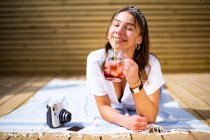 Allegro giovane femmina in abiti casual bere cocktail di frutta fresca e godersi soleggiata giornata estiva mentre si trova sulla terrazza vicino fotocamera istantanea — Foto stock