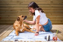 Seitenansicht einer positiven jungen Frau in lässigem Hemd und Jeans-Shorts, die mit einem niedlichen Hund in der Nähe liegt und sich an einem Sommertag auf einer sonnigen Terrasse ausruht — Stockfoto