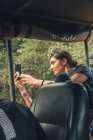 Seitenansicht einer lächelnden Touristin, die während einer Safari im Auto sitzt und Fotos mit dem Smartphone macht — Stockfoto