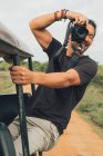 Fotógrafo masculino alegre étnico sentado en el coche y tomando fotos de la naturaleza durante las vacaciones en safari - foto de stock