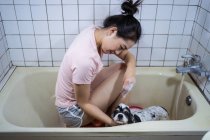 Vista lateral étnica Asiática do sexo feminino proprietário sentado na banheira e lavar bonito Cocker Spaniel filhote de cachorro em casa — Fotografia de Stock