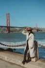 Vista lateral de sorrir jovem senhora na roupa da moda com chapéu e óculos de sol em pé no dique contra Golden Gate Bridge, na Califórnia, em dia ensolarado — Fotografia de Stock