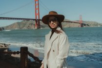 Vista lateral da jovem mulher sorridente em roupa da moda com chapéu e óculos de sol em pé no dique contra a Golden Gate Bridge, na Califórnia, em dia ensolarado — Fotografia de Stock