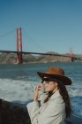 Вид сбоку улыбающейся юной леди в модном наряде в шляпе и солнцезащитных очках, стоящей на набережной у моста Золотые Ворота в Калифорнии в солнечный день — стоковое фото