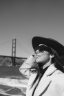 Side view of smiling young lady in trendy outfit with hat and sunglasses standing on embankment against Golden Gate Bridge in California in sunny day — Stock Photo