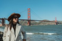 Vista lateral da jovem mulher sorridente em roupa da moda com chapéu e óculos de sol em pé no dique contra a Golden Gate Bridge, na Califórnia, em dia ensolarado — Fotografia de Stock
