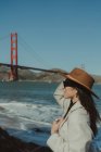 Seitenansicht einer lächelnden jungen Dame im trendigen Outfit mit Hut und Sonnenbrille, die an einem sonnigen Tag auf einem Damm vor der Golden Gate Bridge in Kalifornien steht — Stockfoto