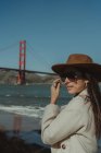 На стіні вимальовується усміхнена молода леді в модному одязі з капелюхом і сонцезахисними окулярами, що стоять на набережній проти моста Золоті Ворота в Каліфорнії в сонячний день. — стокове фото