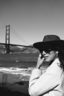 Side view of smiling young lady in trendy outfit with hat and sunglasses standing on embankment against Golden Gate Bridge in California in sunny day — Stock Photo
