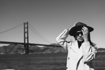 Vue latérale de la jeune femme souriante en tenue tendance avec chapeau et lunettes de soleil debout sur le remblai contre Golden Gate Bridge en Californie par temps ensoleillé — Photo de stock