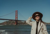 Seitenansicht einer lächelnden jungen Frau im trendigen Outfit mit Hut und Sonnenbrille, die bei sonnigem Wetter auf einer Böschung vor der Golden Gate Bridge in Kalifornien steht — Stockfoto