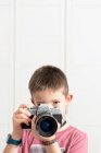 Cute child in casual wear taking photo on retro camera while standing in modern apartment and entertaining during weekend — Stock Photo
