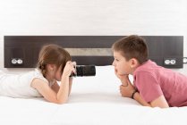 Vista laterale di sorella carina scattare foto di fratello sulla macchina fotografica mentre sdraiato sul letto a casa — Foto stock
